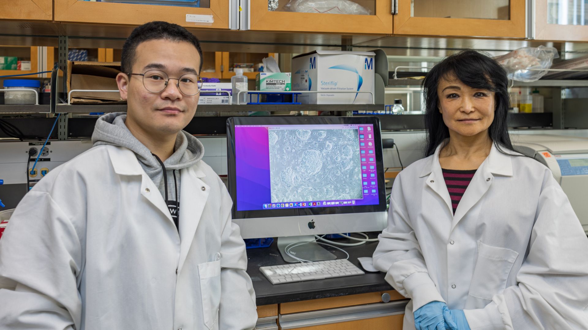 A woman and a man in lab coats posing for a photo in front of a computer screen